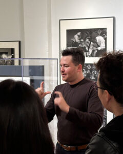 deaf museum educator with fair skin and short brown hair wearing a long sleeve dark shirt is signing "photography" during his tour of images of Billie Holiday at Sugar Hill on view at Woody Guthrie Center