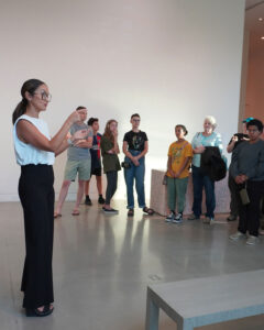 a museum educator wearing black pants and a white sleeveless blouse signs to a diverse group of interested patrons to the arts
