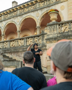 a Deaf museum educator with brown skin wearing a black blouse and trousers stands on the art deco steps of the Philbrook Museum of Art, as patrons look up at her signing about the history of the grounds